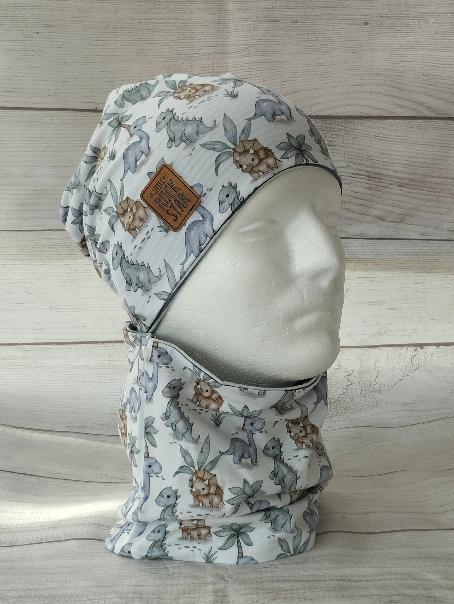 Kindermütze und Loop im Dino-Design auf einem Mannequin – aus weichem Jersey mit Dinosauriern und Pflanzen in gedeckten Farben. Beanie mit Patch ‚Little Rock Sar‘ vor hellem Holz-Hintergrund.