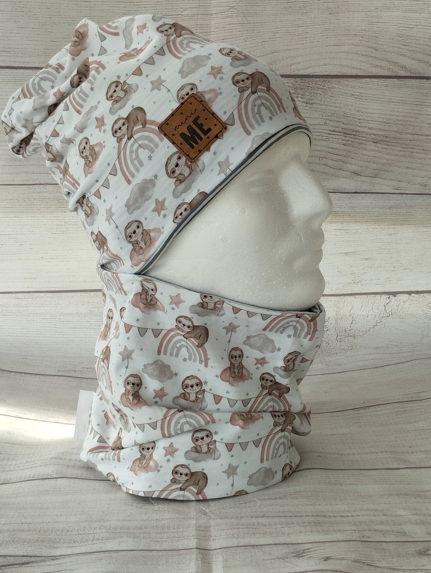 Kindermütze und Loop im Faultier-Design auf einem Mannequin – aus weißem Jersey mit pastellfarbenen Illustrationen von Faultieren, Regenbögen, Sternen und Wolken. Beanie mit Patch ‚MINI ME‘ vor hellem Holz-Hintergrund