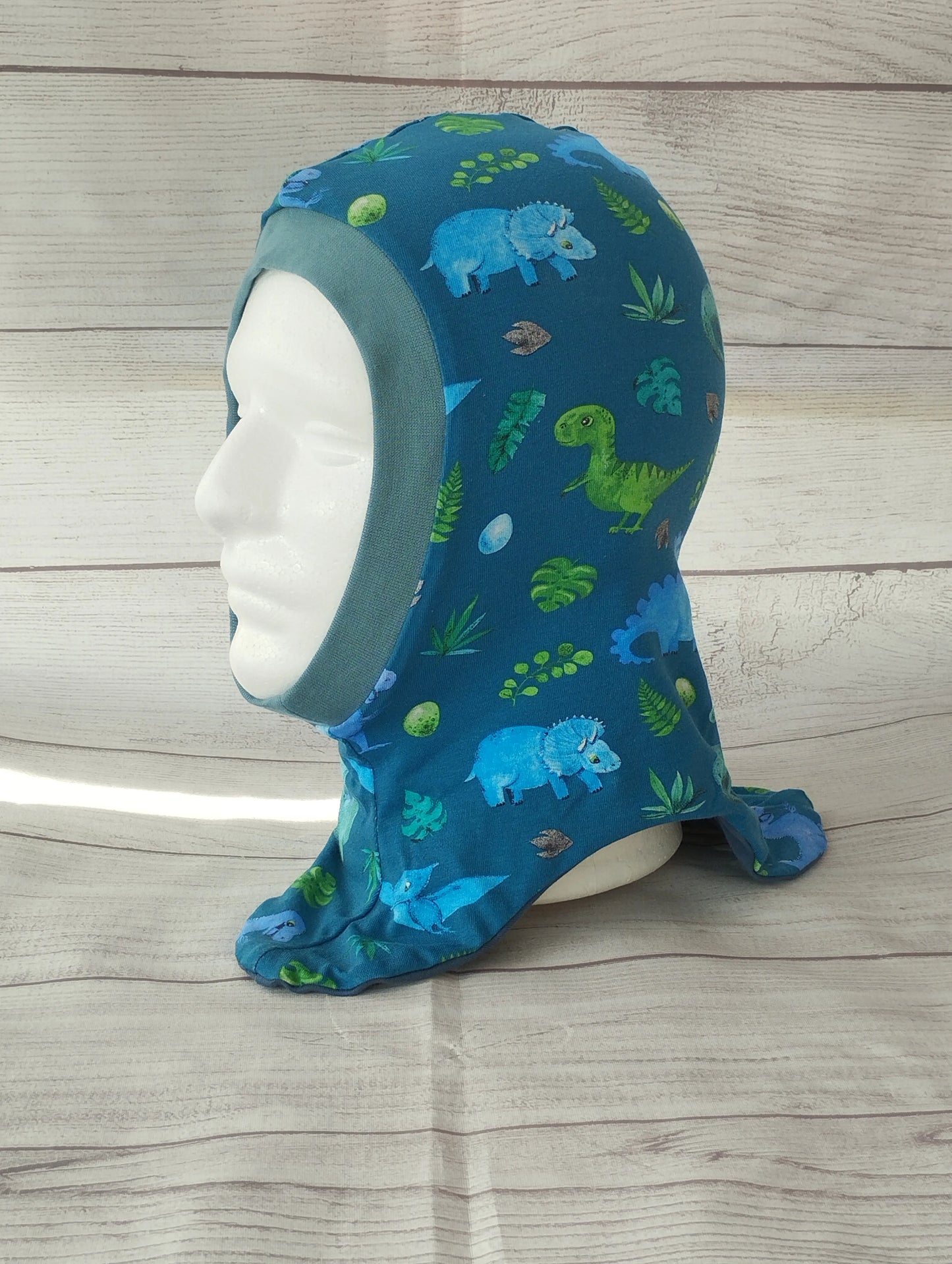 Kinderschlupfmütze im Dino-Design auf einem Mannequin – aus blauem Jersey mit bunten Illustrationen von Dinosauriern, Blättern und Eiern. Präsentiert vor hellem Holz-Hintergrund.