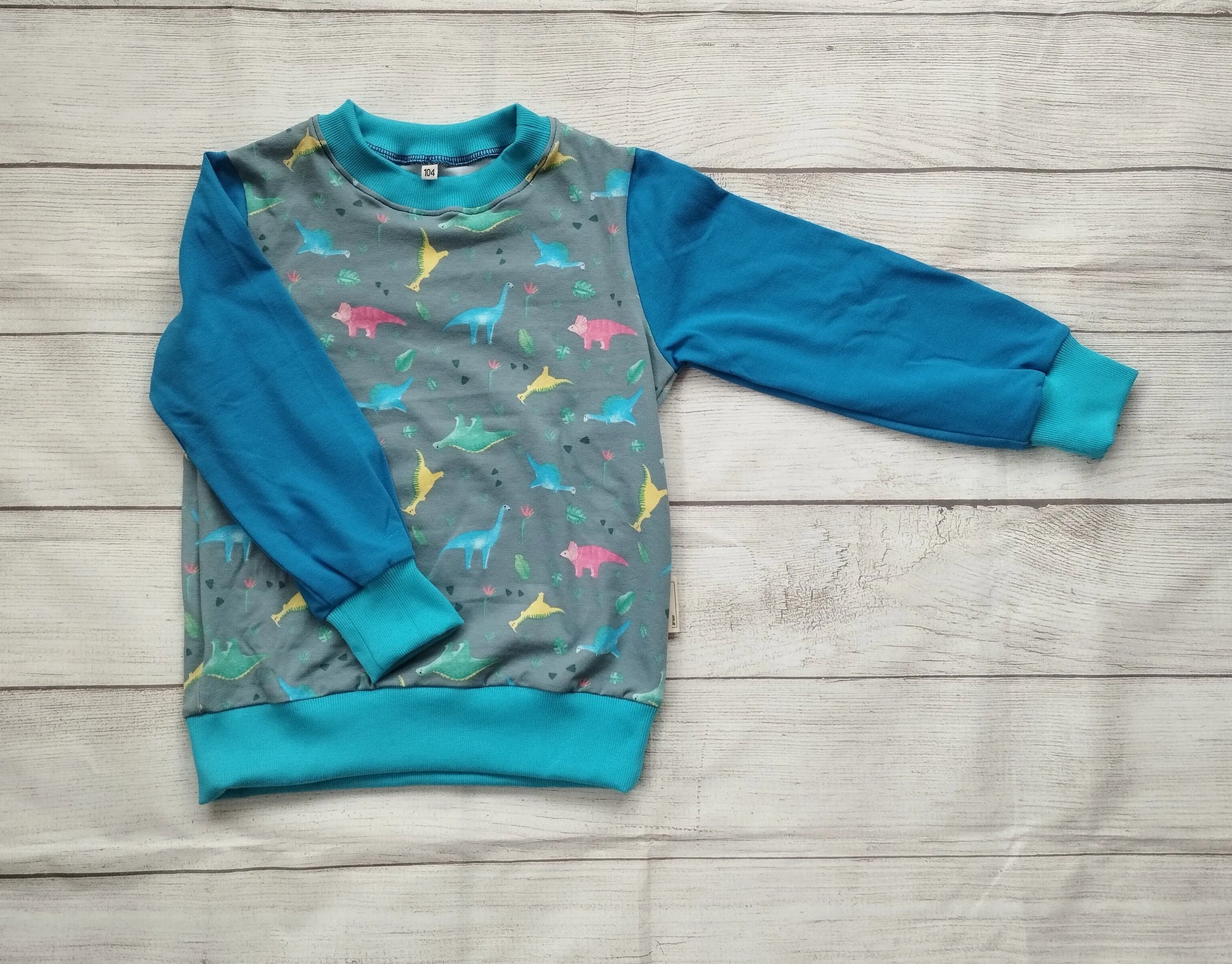 Kinderpullover mit buntem Dino-Muster auf grauem Stoff und blauen Raglanärmeln – handgemacht aus elastischem Baumwolljersey