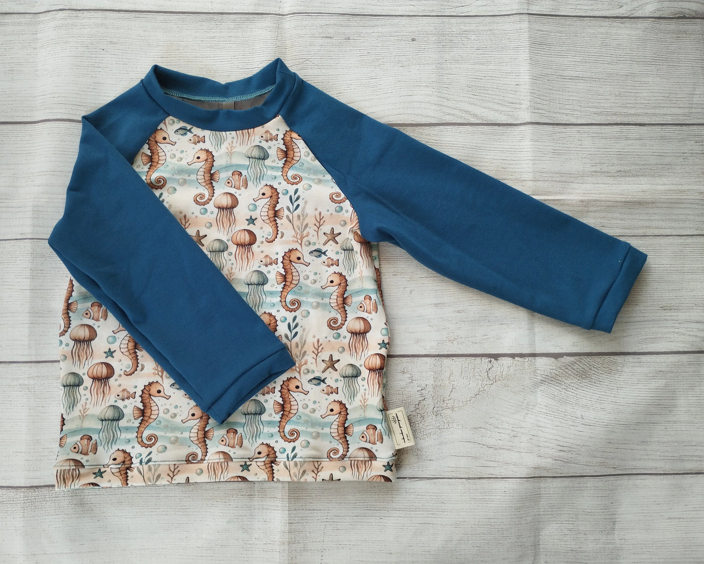 Kinder-Raglanshirt mit Meeresmuster aus Seepferdchen, Quallen und Korallen auf hellem Stoff – blaue Ärmel, handgemacht aus Baumwolljersey