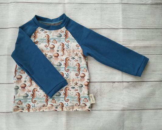 Kinder-Raglanshirt mit Meeresmuster aus Seepferdchen, Quallen und Korallen auf hellem Stoff – blaue Ärmel, handgemacht aus Baumwolljersey