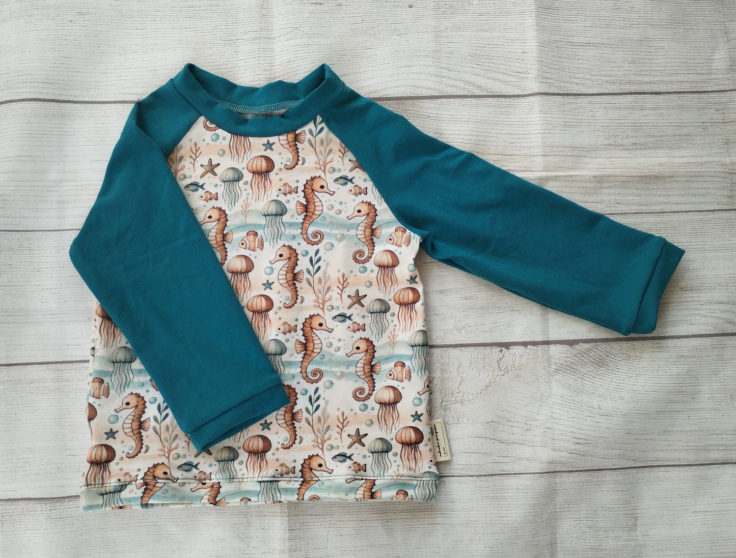 Kinder-Raglanshirt mit Meeresmuster aus Seepferdchen, Quallen und Korallen auf hellem Stoff – petrolfarbene Ärmel, handgemacht aus Baumwolljersey