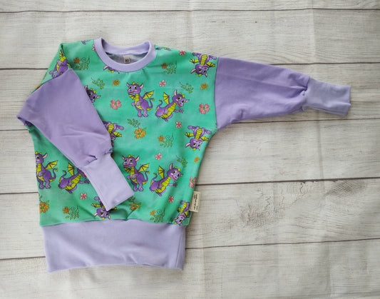 Langarmshirt für Kinder mit lila Ärmeln und grünem Frontpanel – verspieltes Drachenmotiv mit Blumen und Schmetterlingen, handgemacht aus Baumwolljersey