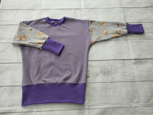 Mitwachspullover für Babys mit lila Bündchen und liebevoller Farbkomposition – flach auf Holzoberfläche präsentiert
