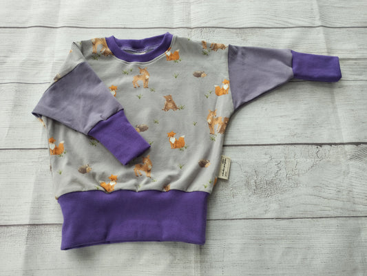 Mitwachspullover für Babys mit Reh, Fuchs und Eichhörnchen auf grauem Stoff – flach auf Holzoberfläche präsentiert