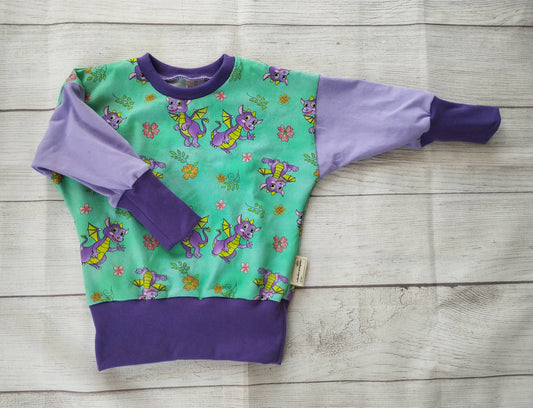 Kinderpullover mit asymmetrischen lila Ärmeln und grünem Vorderteil – verspieltes Drachenmotiv mit Sternen und Blumen, handgemacht aus Baumwolljersey