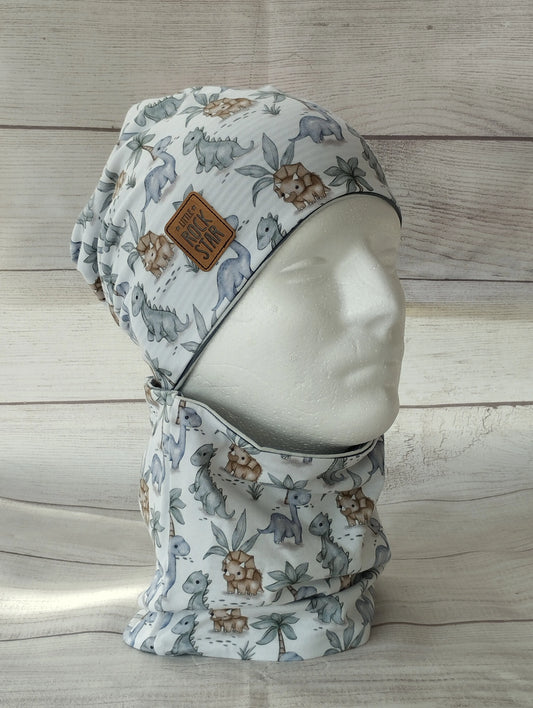 Kindermütze und Loop im Dino-Design auf einem Mannequin – aus weichem Jersey mit Dinosauriern und Pflanzen in gedeckten Farben. Beanie mit Patch ‚Stay Wild‘ vor hellem Holz-Hintergrund.