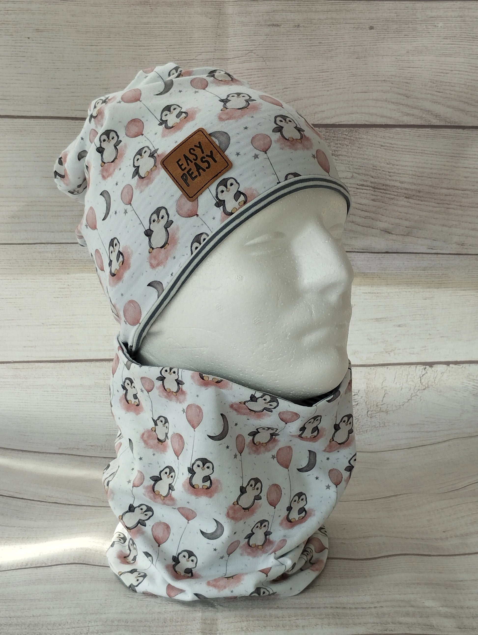 Kindermütze und Loop im Pinguin-Ballon-Design auf einem Mannequin – aus weißem Jersey mit rosa Akzenten, verziert mit Pinguinen, Ballons, Monden und Sternen. Präsentiert vor hellem Holz-Hintergrund