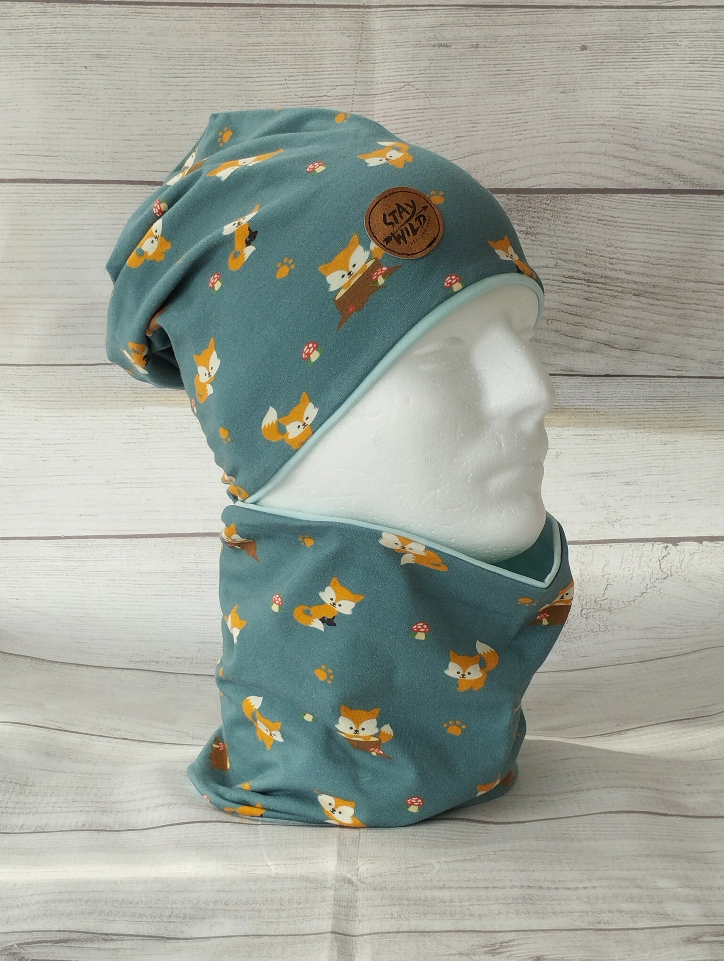 Kindermütze und Loop im Set auf einem Mannequin – aus petrolfarbenem Jersey mit Fuchs-Motiv. Handgemachte Winteraccessoires vor rustikalem Holz-Hintergrund.