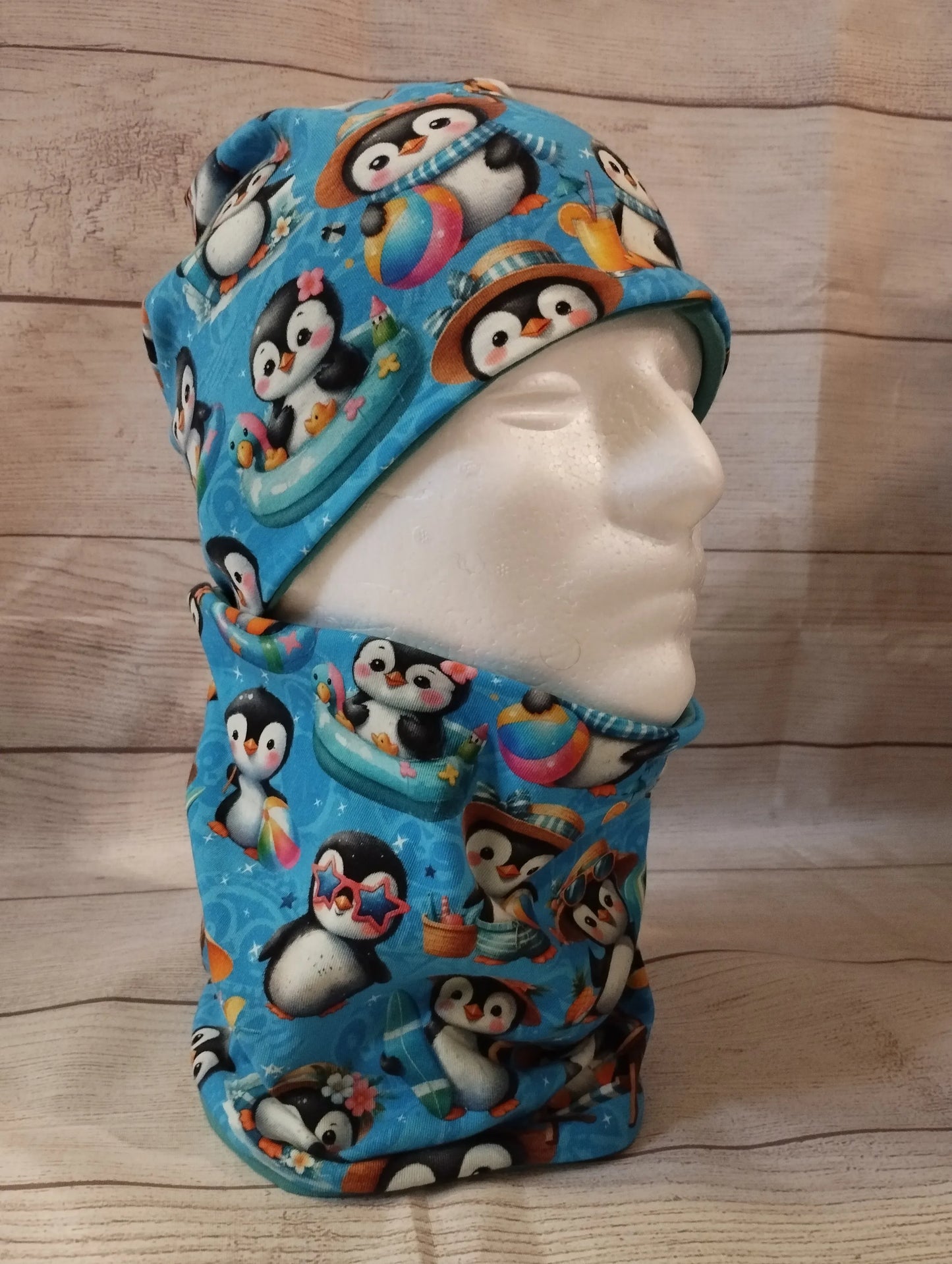 Kindermütze und Loop im sommerlichen Pinguin-Design auf einem Mannequin – aus blauem Jersey mit fröhlichen Illustrationen von Pinguinen beim Baden, Spielen und Sonnen. Präsentiert vor hellem Holz-Hintergrund.
