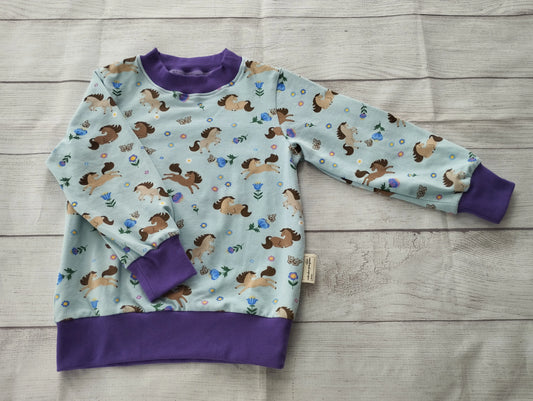 Kinderpullover mit springenden Pferden auf hellblauem Stoff und lila Bündchen – handgemacht aus elastischem Baumwolljersey