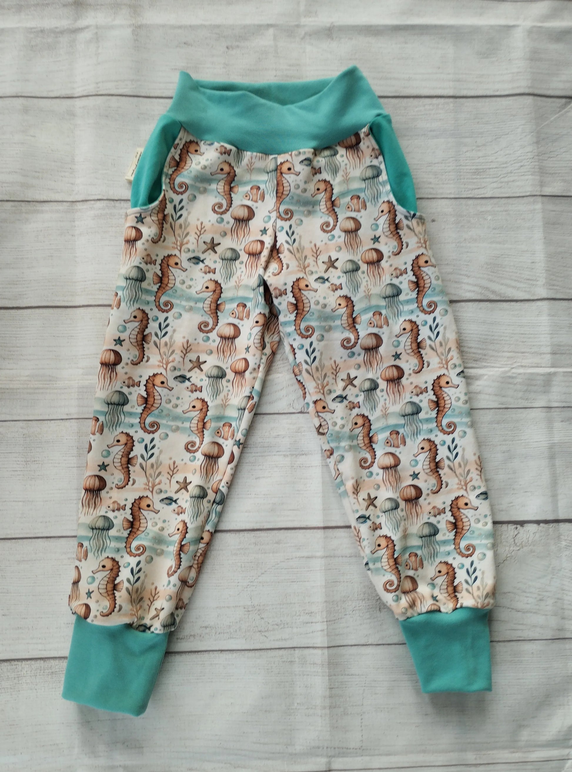 Handgemachte Pumphose mit Seepferdchen- und Meeresmotiv, flach ausgelegt auf hellem Holz – elastischer Baumwolljersey mit Bündchen in Türkis.