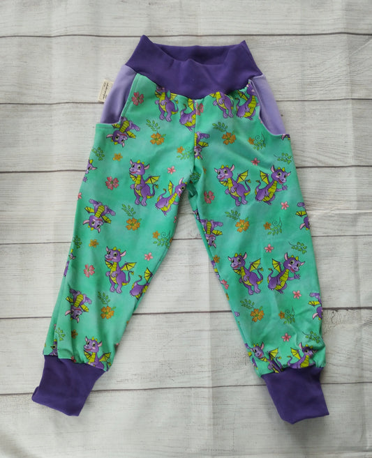 Handgemachte Pumphose mit lila Drachen auf mintfarbenem Grund, flach ausgelegt auf Holz – elastischer Baumwolljersey mit lila Bündchen und Taschen.