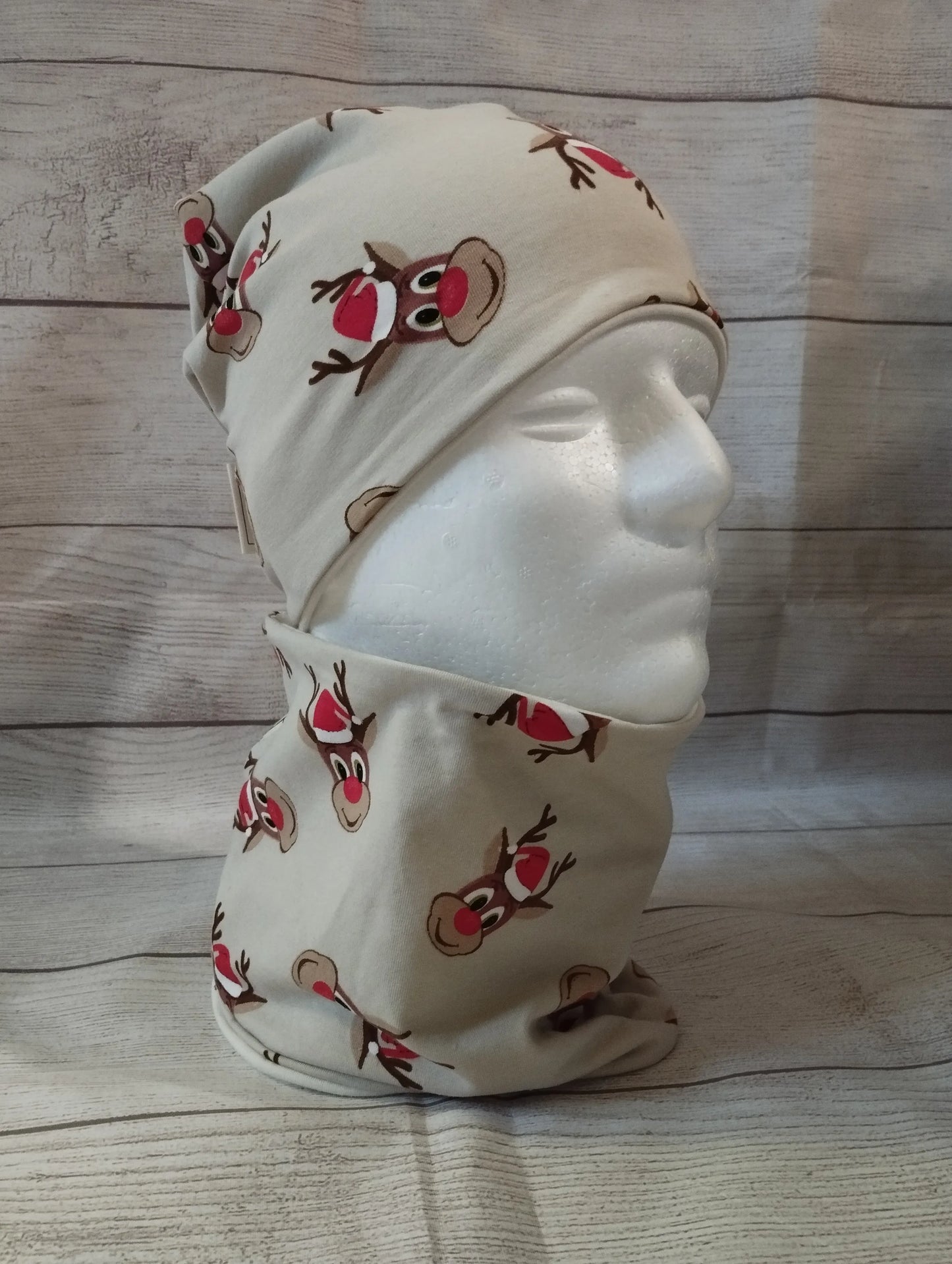 Kindermütze und Loop im Rentier-Design auf einem Mannequin – aus beigefarbenem Jersey mit roten Rentiernasen und festlichen Illustrationen. Präsentiert vor rustikalem Holz-Hintergrund.