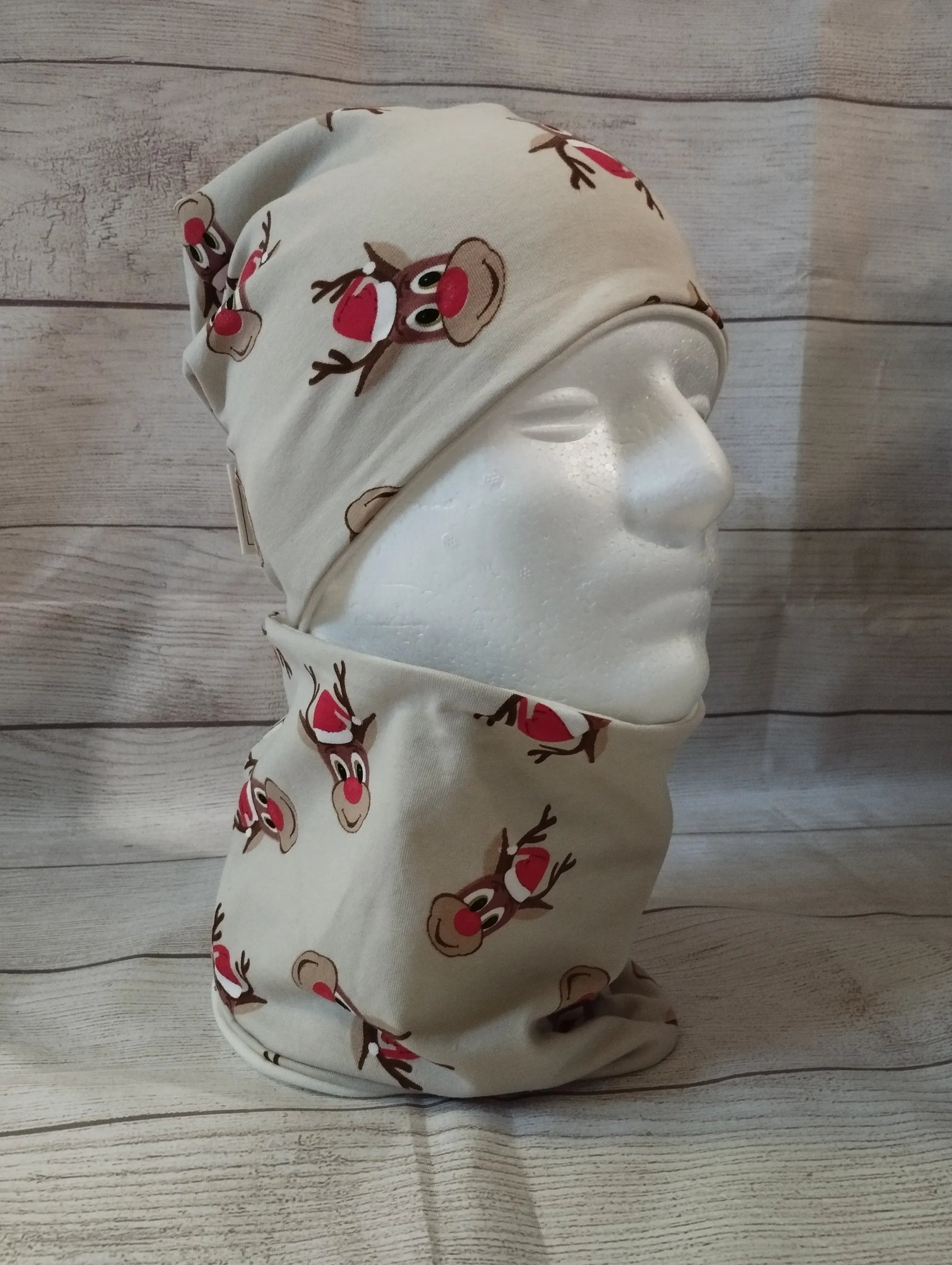 Kindermütze und Loop im Rentier-Design auf einem Mannequin – aus beigefarbenem Jersey mit roten Rentiernasen und festlichen Illustrationen. Präsentiert vor rustikalem Holz-Hintergrund.