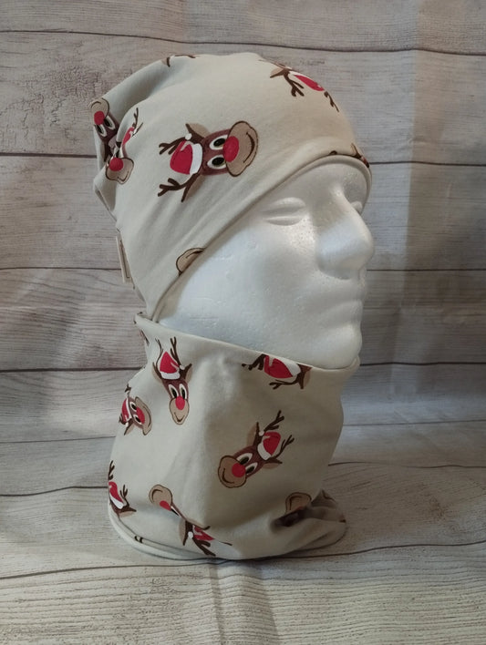 Kindermütze und Loop im Rentier-Design auf einem Mannequin – aus beigefarbenem Jersey mit roten Rentiernasen und festlichen Illustrationen. Präsentiert vor rustikalem Holz-Hintergrund.