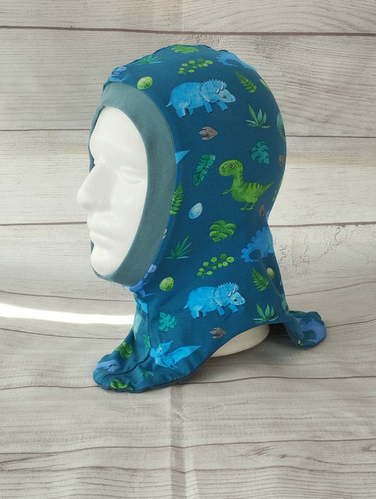 Kinderschlupfmütze im Dino-Design auf einem Mannequin – aus blauem Jersey mit bunten Illustrationen von Dinosauriern, Blättern und Eiern. Präsentiert vor hellem Holz-Hintergrund.