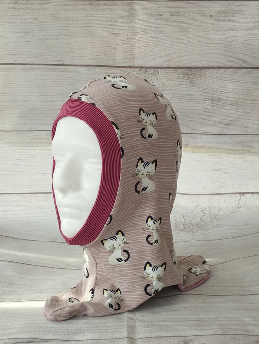 Kinderschlupfmütze im Katzen-Design auf einem Mannequin – aus rosa Jersey mit niedlichen Katzenköpfen, geripptem dunkelrosa Bündchen am Gesichtsausschnitt. Präsentiert vor hellem Holz-Hintergrund.