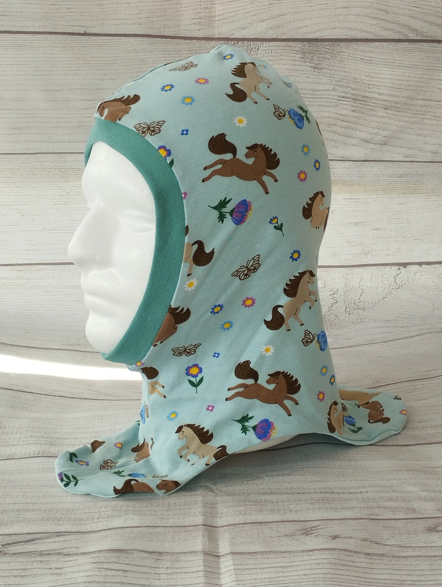 Kinderschlupfmütze im Pferde-Design auf einem Mannequin – aus hellblauem Jersey mit bunten Illustrationen von Pferden, Schmetterlingen, Blumen und Eicheln. Gesichtsausschnitt mit türkisfarbenem Bündchen eingefasst, präsentiert vor hellem Holz-Hintergrund.