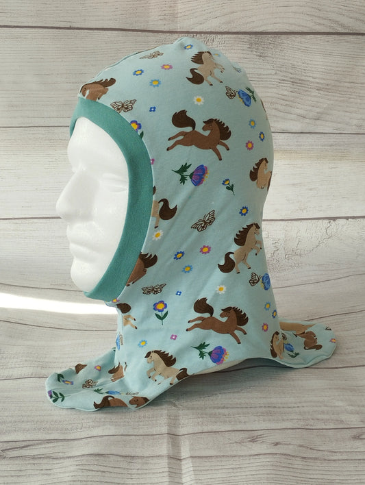Kinderschlupfmütze im Pferde-Design auf einem Mannequin – aus hellblauem Jersey mit bunten Illustrationen von Pferden, Schmetterlingen, Blumen und Eicheln. Gesichtsausschnitt mit türkisfarbenem Bündchen eingefasst, präsentiert vor hellem Holz-Hintergrund.