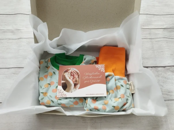Geschenkbox mit handgemachter Babykleidung in Pfirsichmuster, liebevoll verpackt in weißen Seidenpapier, mit Grußkarte zur Geburt 

